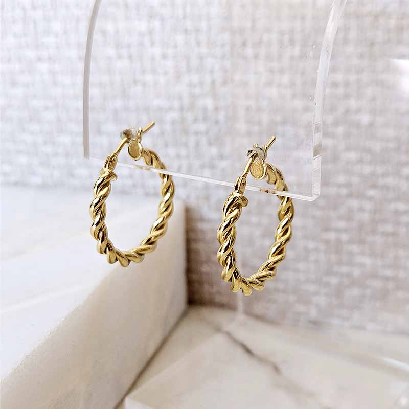 A pair of gold twisted hoop earrings hanging from a clear acrylic display stand against a soft beige textured background.