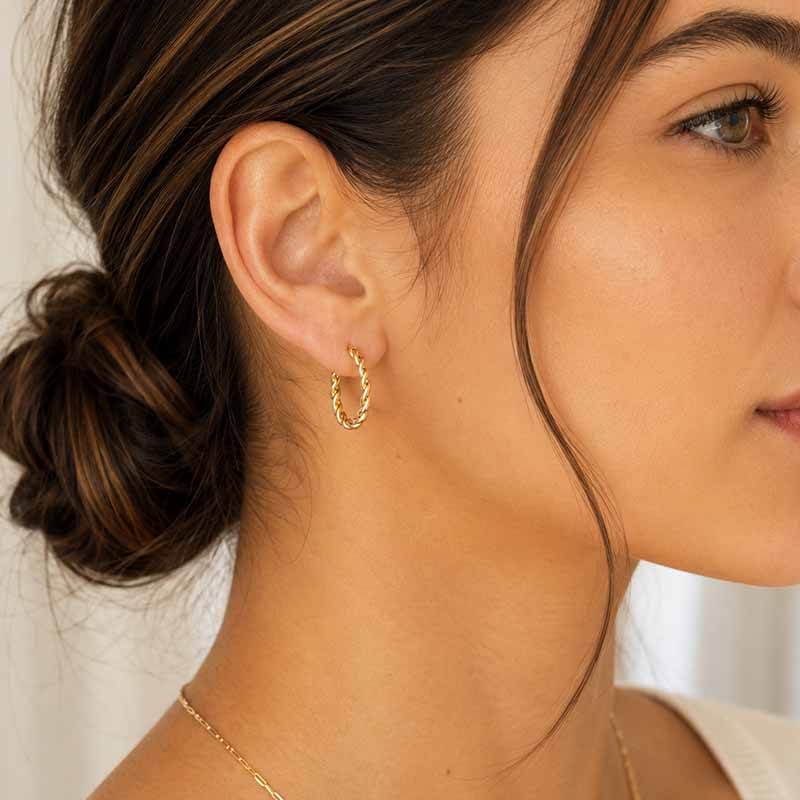 A close-up of a woman wearing a delicate gold hoop earring with a twisted, textured finish. The shot focuses on her ear and side profile, showing the jewelry in a lifestyle context.