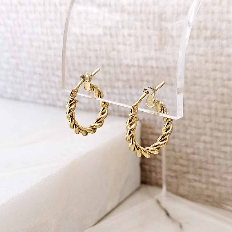 A pair of gold hoop earrings with a twisted rope texture, displayed on a clear acrylic stand against a neutral beige textured background.