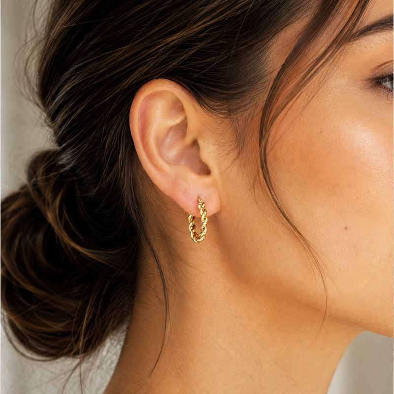 A close-up of a woman's ear wearing a small twisted gold hoop earring.