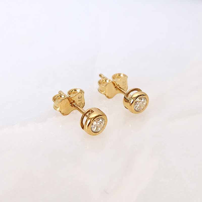 A pair of gold stud earrings with round clear gemstones, shown on a light off-white marble surface.