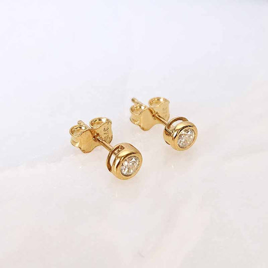 A pair of gold stud earrings with round clear gemstones, shown on a light off-white marble surface.