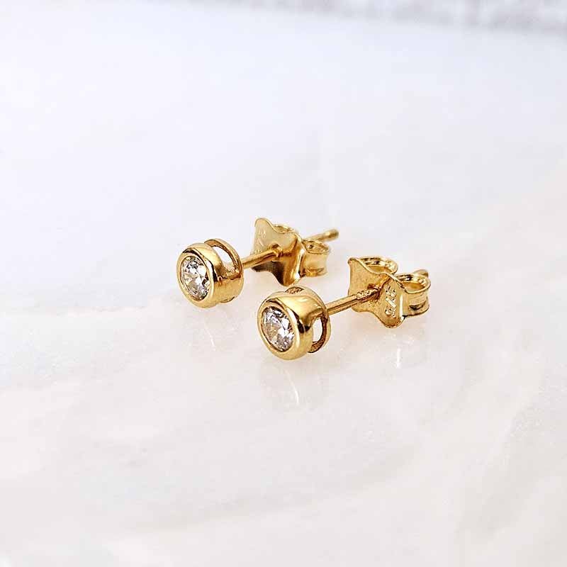 A pair of gold stud earrings with round bezel-set clear gemstones, displayed on a light, reflective marble surface.
