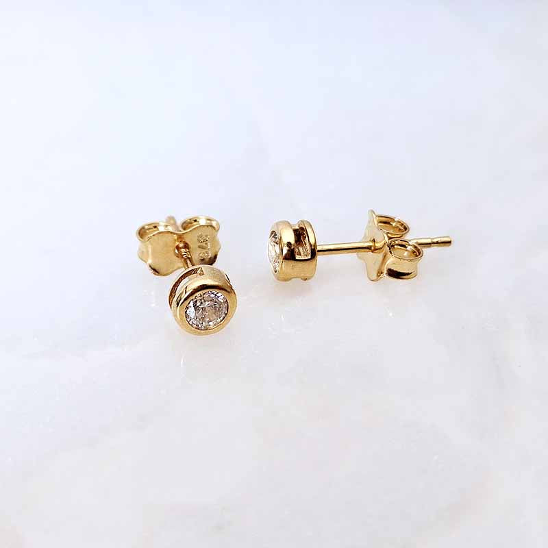 A pair of gold stud earrings with round bezel-set clear gemstones, shown on a light white marble background. One earring is facing forward, and the other is turned to show the side profile.
