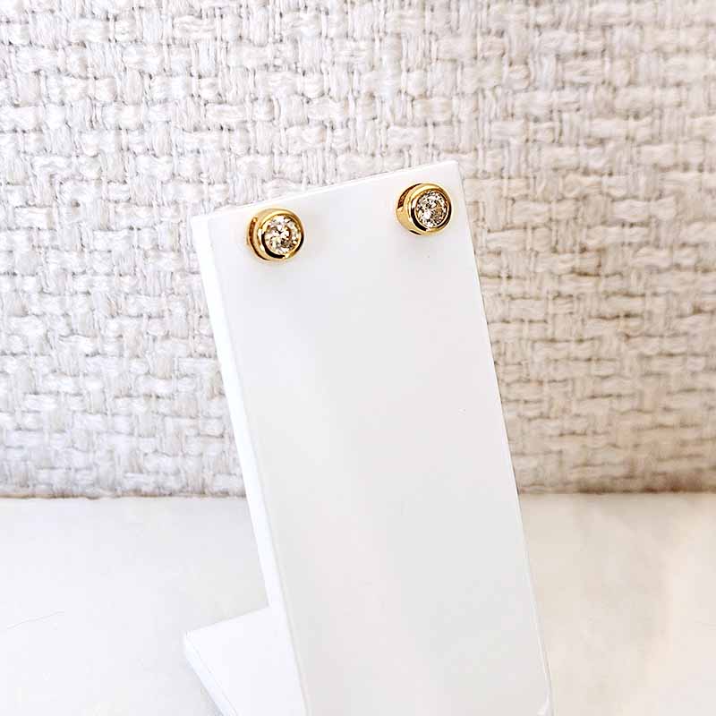 A pair of gold stud earrings with clear gemstones, displayed on a white stand against a textured beige woven background.
