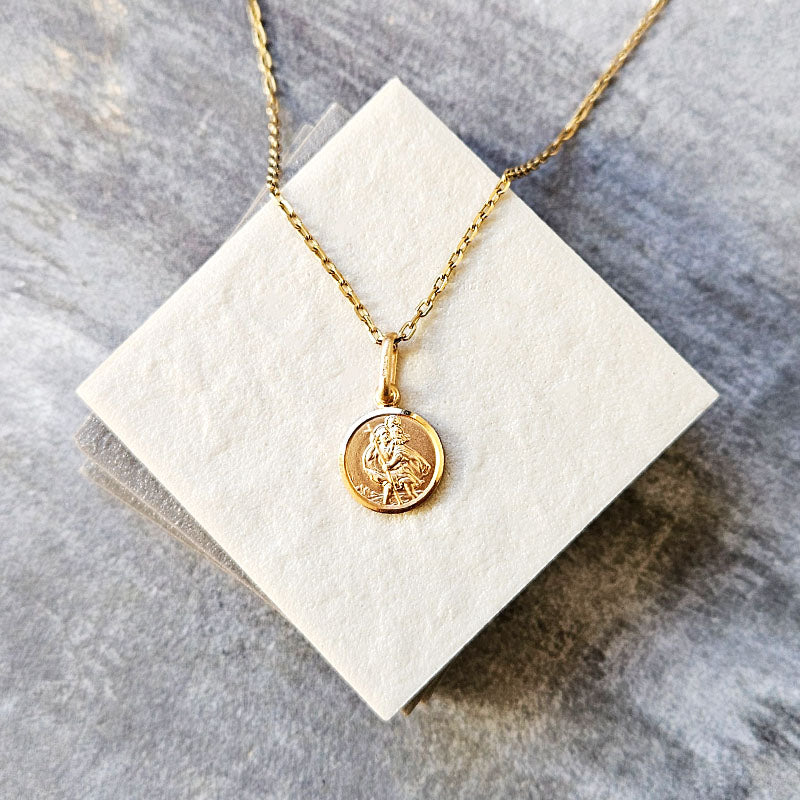 Gold necklace with a St Christopher pendant on a white square card against a gray marble background
