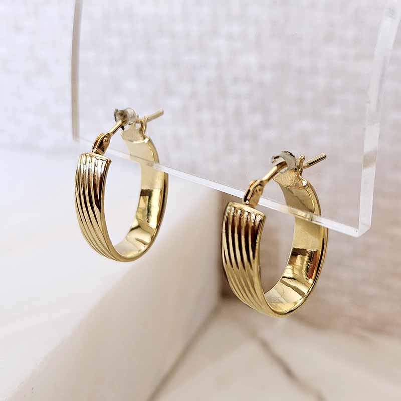 A pair of gold hoop earrings with a textured, ridged outer band, displayed on a clear acrylic stand against a light beige background.