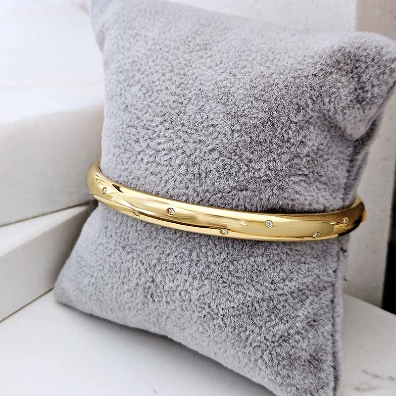 A polished gold bangle bracelet with small embedded clear stones, displayed on a textured gray plush pillow.