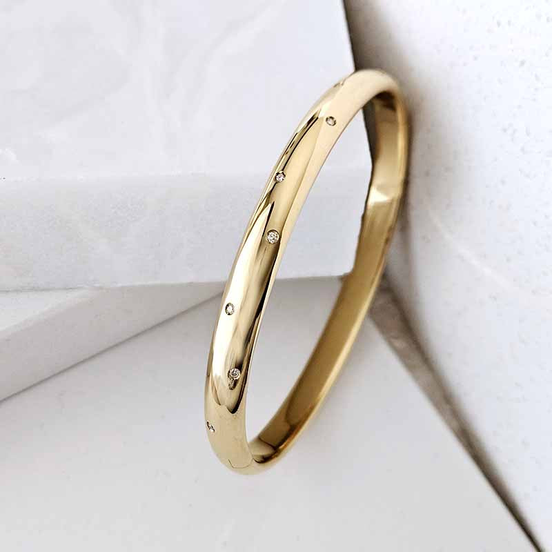A polished gold bangle bracelet with small evenly spaced clear gemstones, photographed on a clean white textured surface.
