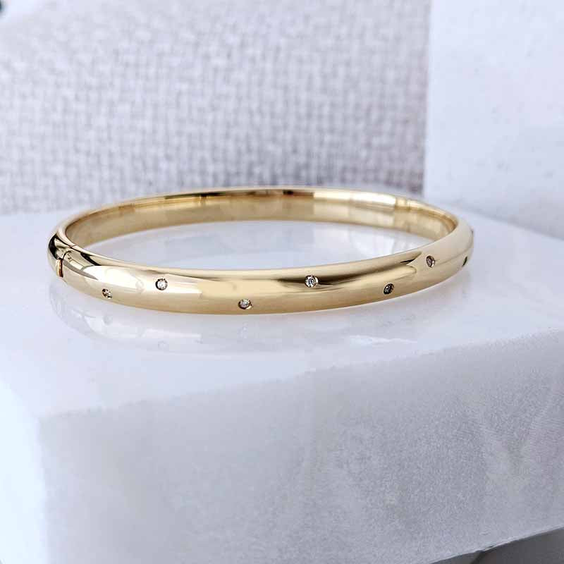 A polished gold bangle bracelet with small inset clear stones spaced along the band, displayed on a white textured surface.