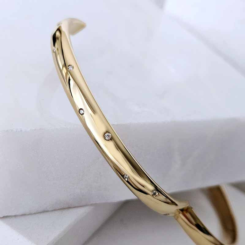 A polished gold bangle bracelet with small inset clear diamonds, displayed on a white textured surface.