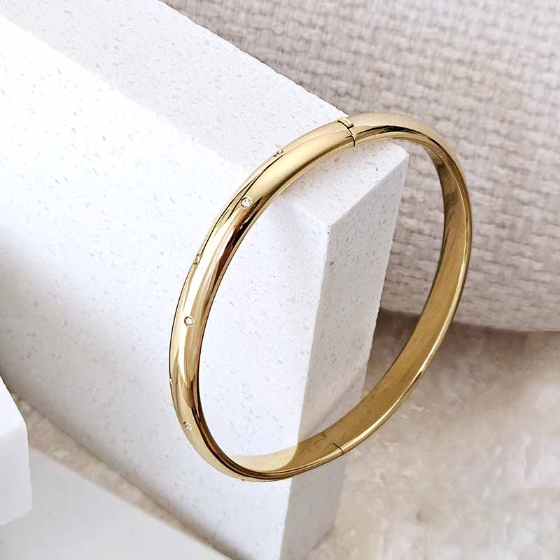 A polished gold bangle bracelet with tiny inset stones, resting on a white textured display block.