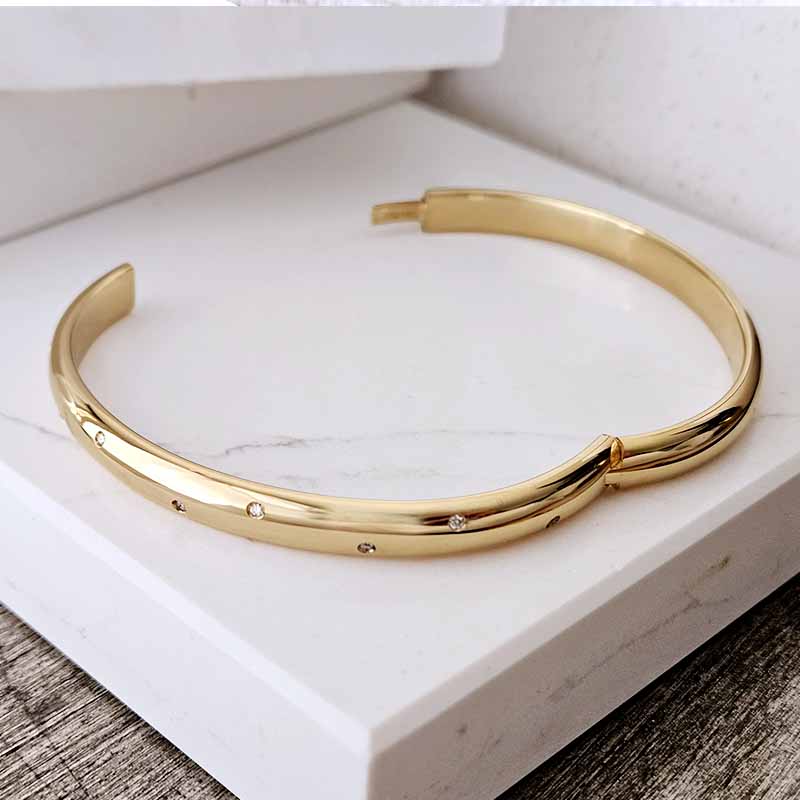 A polished gold-toned open cuff bangle bracelet with small inset clear stones, displayed on a white marble block.
