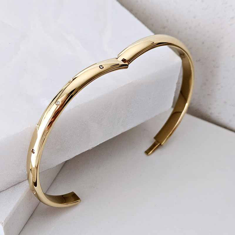 A polished gold-tone open cuff bracelet with small engraved geometric details, displayed on a white textured surface.