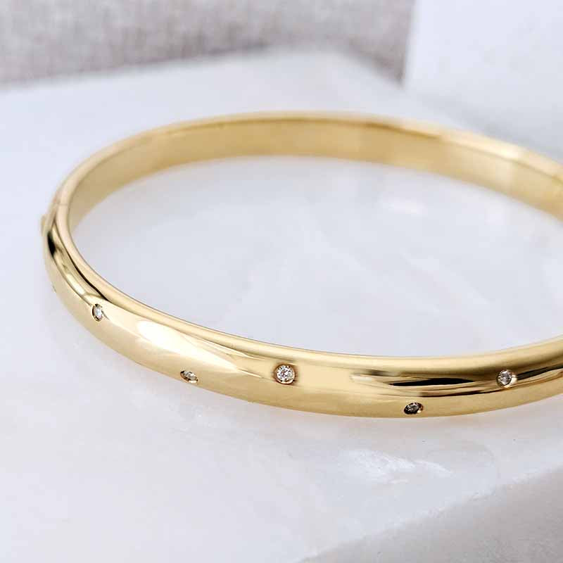 A polished yellow gold bangle bracelet with small inset clear stones spaced along the band, photographed close-up on a light marble-like surface.