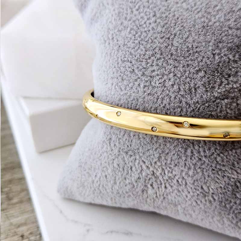A polished gold bangle bracelet with small inset stones, displayed on a textured gray cushion.