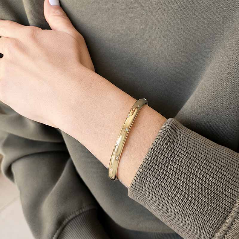 A close-up of a person's wrist wearing a polished gold bangle bracelet, shown against a dark olive green ribbed sweatshirt.