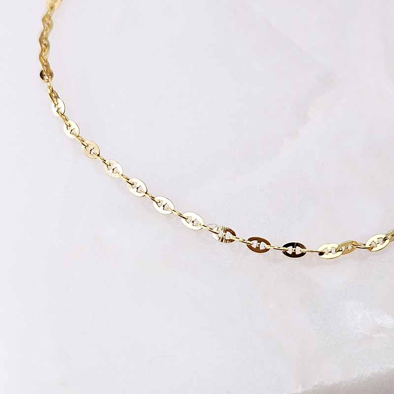 A close-up of a delicate gold chain bracelet featuring alternating flat oval links and small round links, laid out on a light, off-white textured surface.