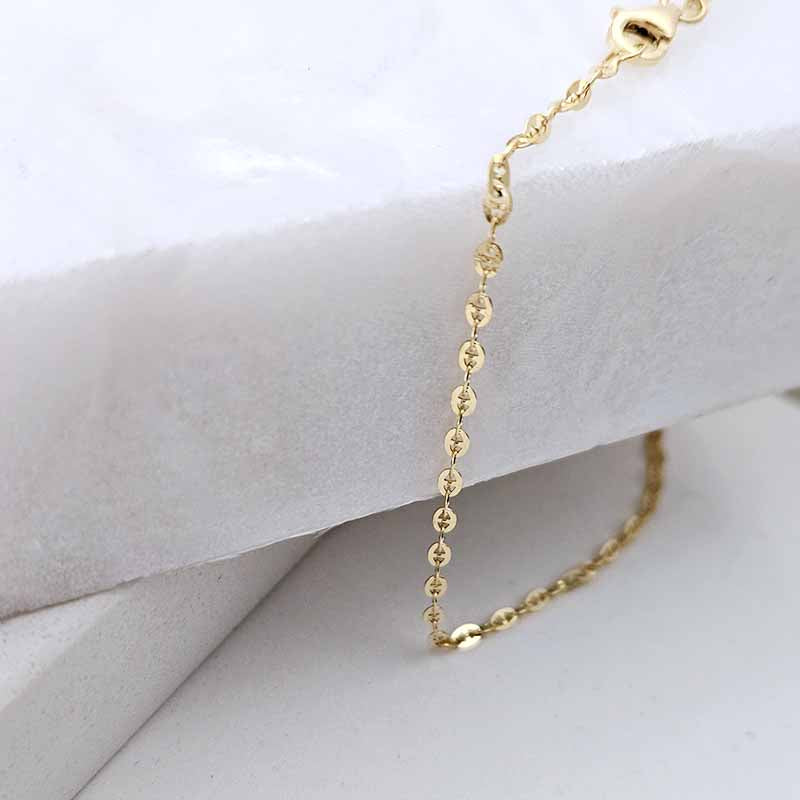 A close-up of a delicate gold chain necklace draped over a white textured display block, showing the fine links and clasp.