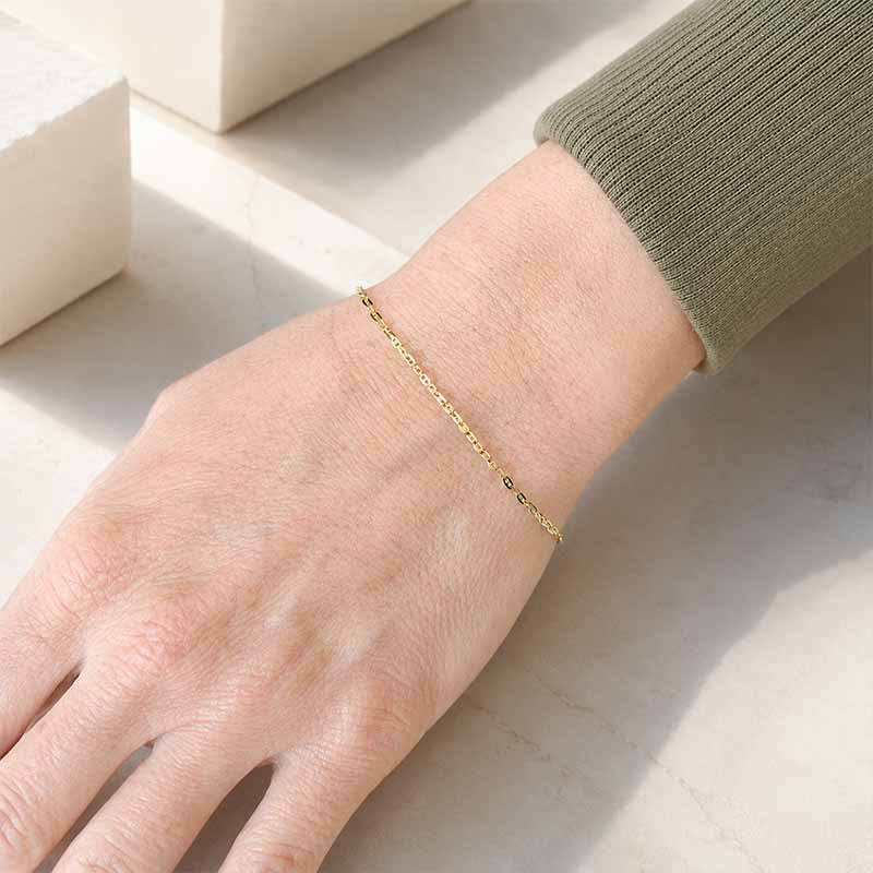A delicate gold chain bracelet worn on a person's wrist, shown in a close-up shot with a light olive green ribbed sweater sleeve visible.