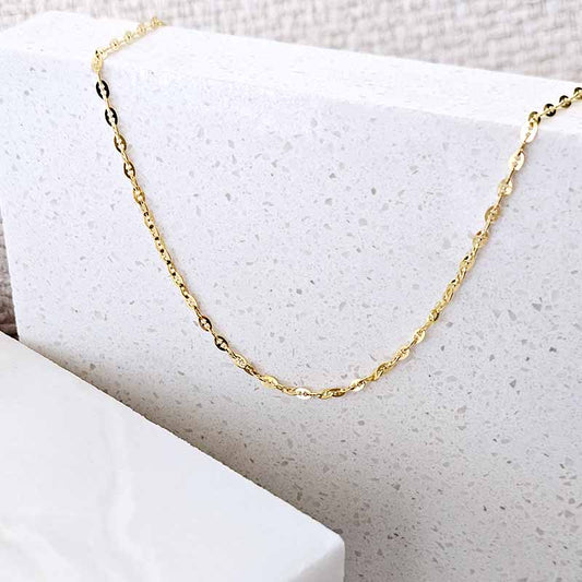 A delicate gold chain necklace draped over a textured white stone block, showcasing its fine links and polished finish.