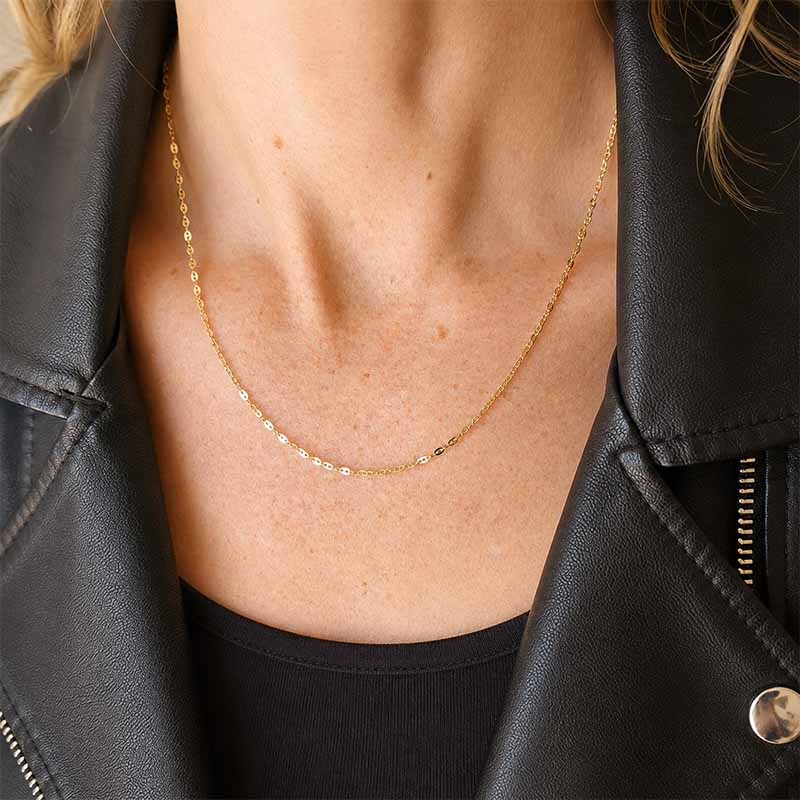 A delicate gold chain necklace with small clear stones, worn by a model in a black leather jacket over a black top.
