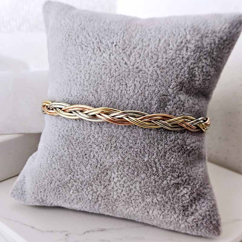 A braided cable knot bracelet in mixed gold and rose gold metals, displayed on a textured gray velvet pillow.