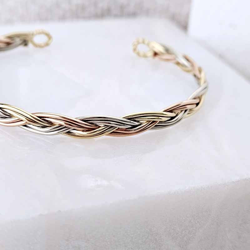 A close-up of a braided cuff bracelet with interwoven gold, silver, and rose gold metal strands, resting on a white textured surface.