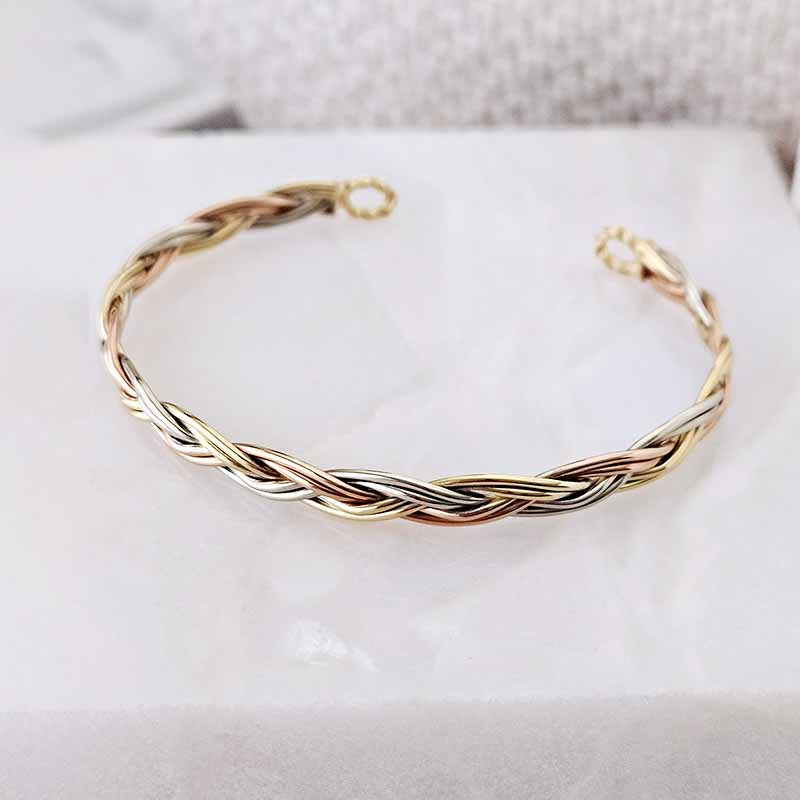 A twisted braided cuff bracelet in a two-tone gold and silver finish, displayed on a white textured surface.