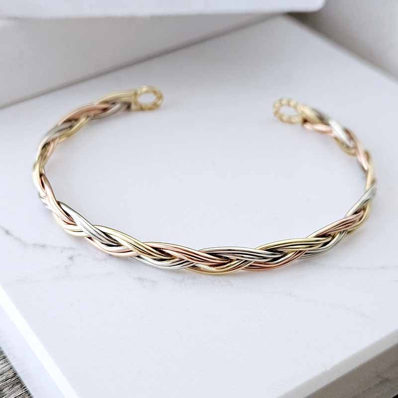 A twisted three-metal cable cuff bracelet in gold, rose gold, and silver, displayed on a white marble surface.