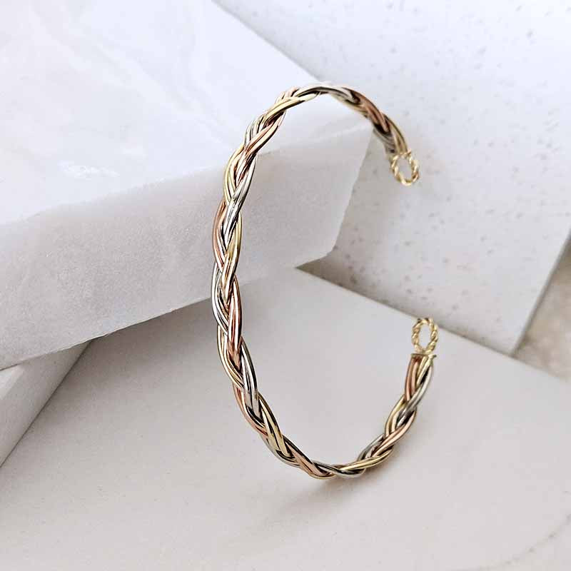 A twisted braided cuff bracelet in a three-tone gold, silver, and rose gold finish, displayed on a white marble block.