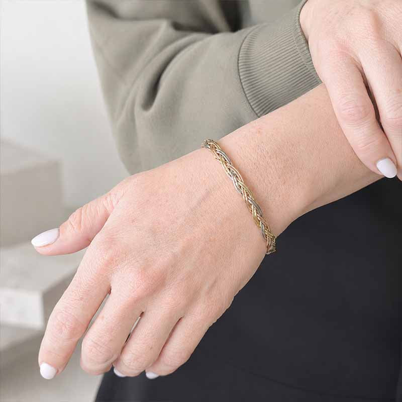 A close-up of a person's wrist wearing a delicate braided bracelet in gold and silver tones, paired with a taupe long-sleeve top and black pants.