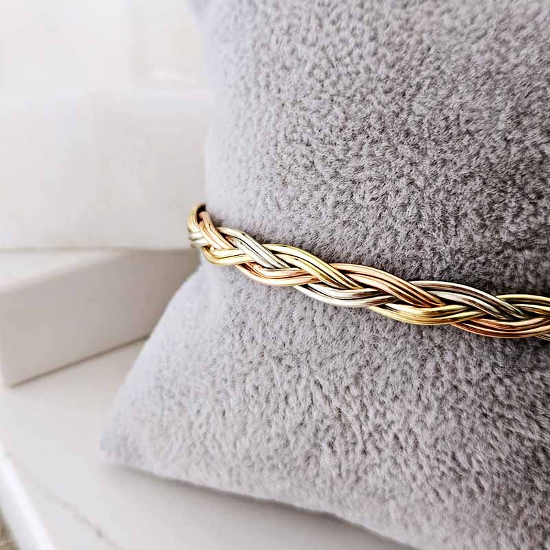 A close-up of a braided metallic bracelet in gold and silver tones, wrapped around a soft gray textured pillow.