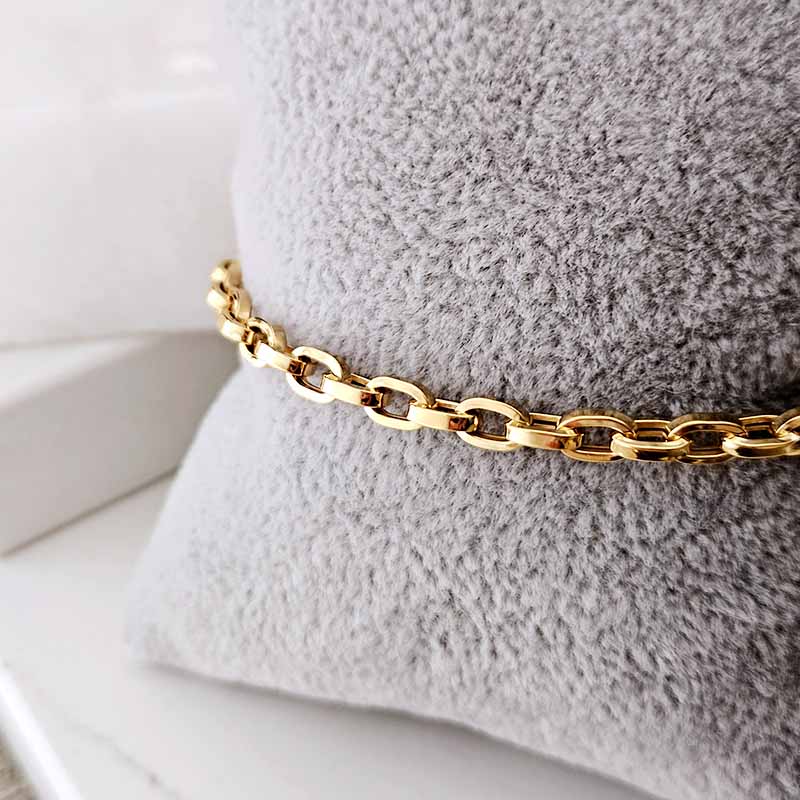 A close-up of a polished gold chain bracelet resting on a textured gray display cushion.