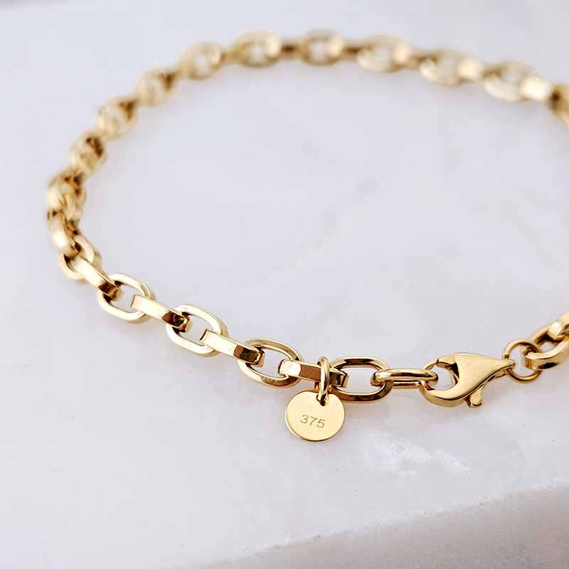 A gold chain bracelet with a lobster clasp and a small round tag engraved with '375', displayed on a light marble surface.