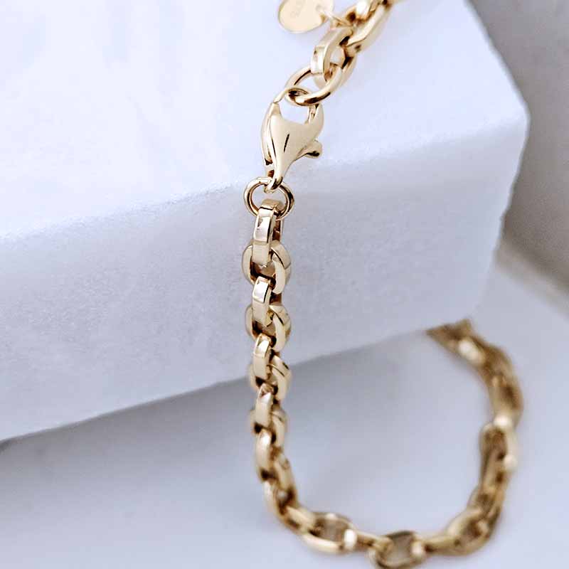 A close-up of a polished gold chain bracelet draped over a white jewelry display block, showing the lobster clasp and interlocking links.