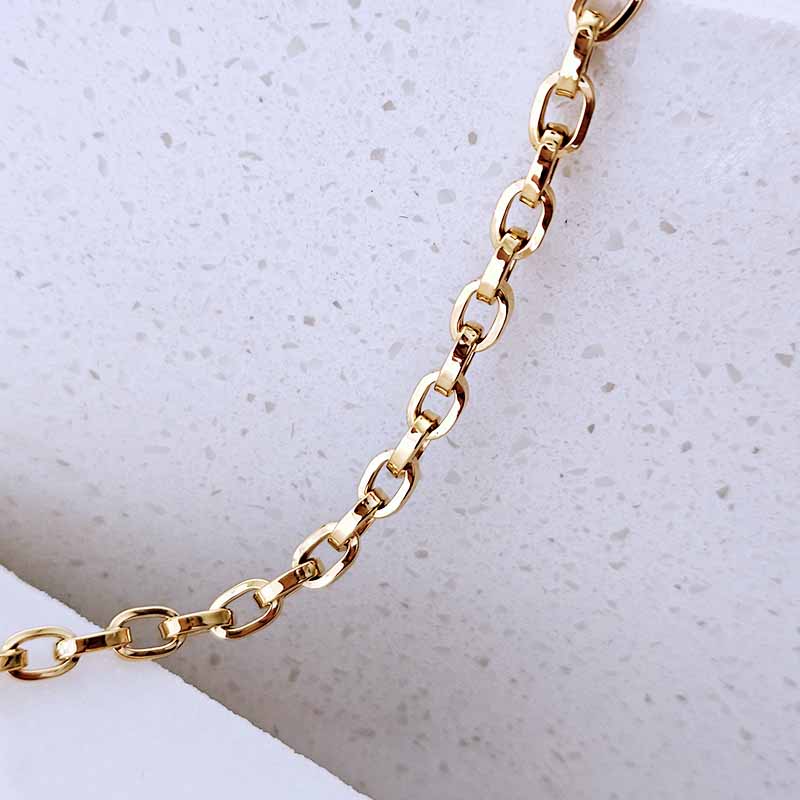 A close-up of a polished gold chain with elongated oval links, laid diagonally across a textured white surface.