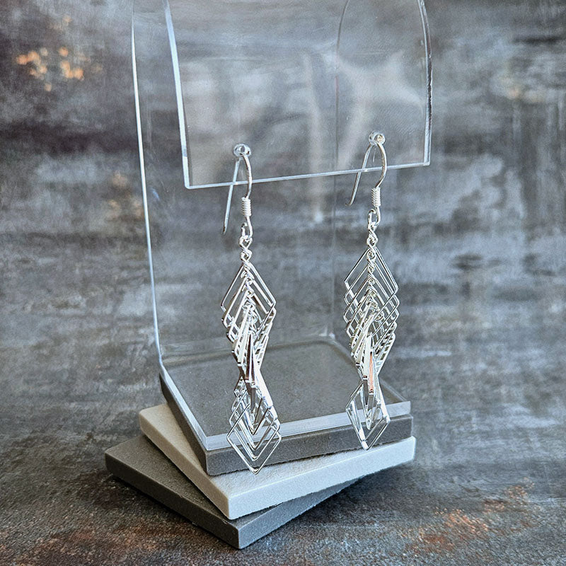 Silver geometric earrings on a clear stand against a textured gray background