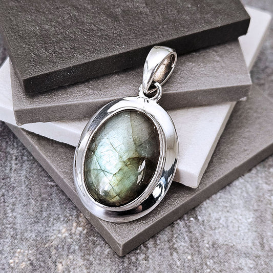 A silver oval pendant featuring a greenish-gray labradorite gemstone, displayed on stacked gray stone blocks.