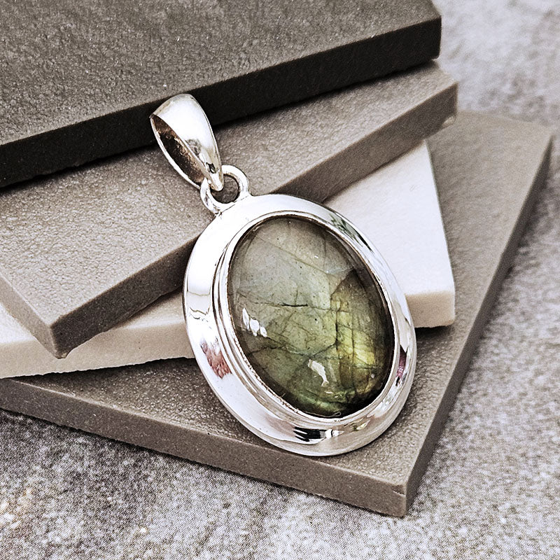 A silver oval pendant featuring a greenish-gray labradorite gemstone with natural crystalline patterns, displayed on stacked gray stone blocks.