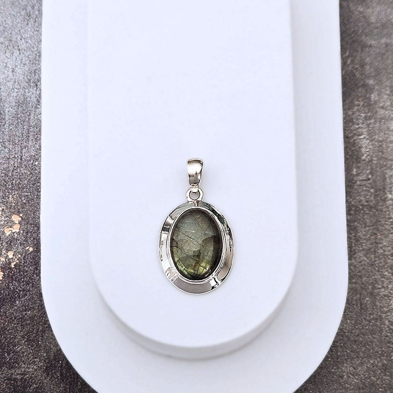 A silver oval pendant featuring a greenish-gray labradorite stone, displayed on a white curved stand against a textured gray background.