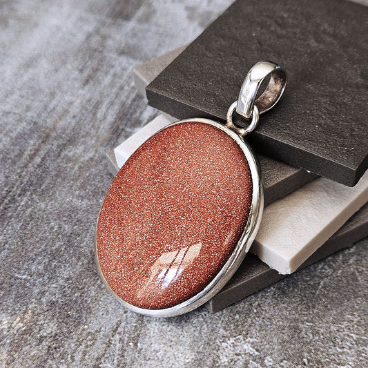 A silver bezel-set oval pendant featuring a shimmering coppery-brown glitter stone, displayed on stacked dark and light gray stone blocks.