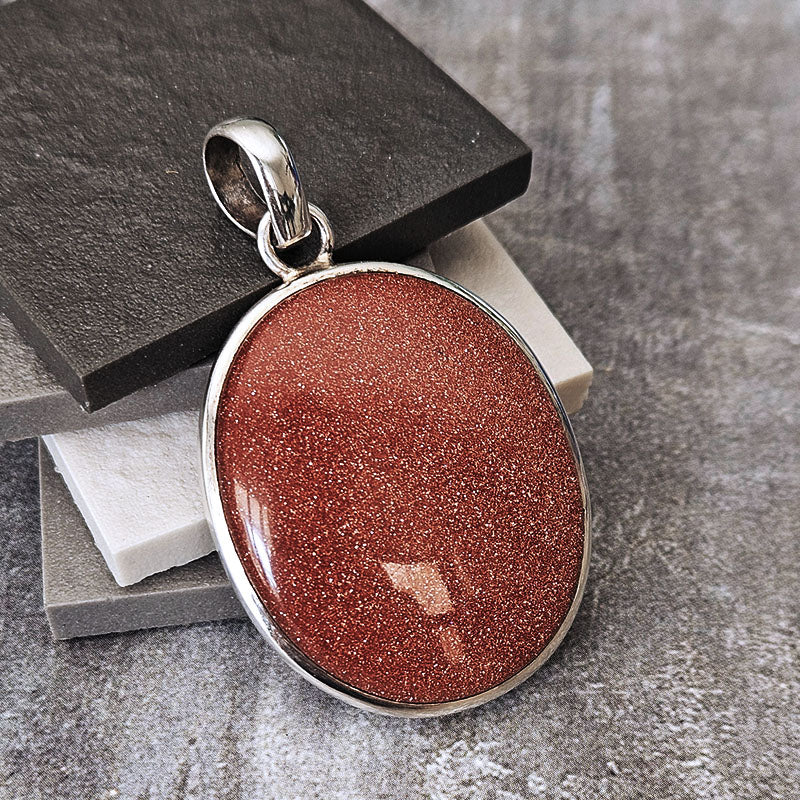 An oval pendant featuring a sparkling coppery red stone in a polished silver bezel, resting on stacked dark and light gray stone slabs.