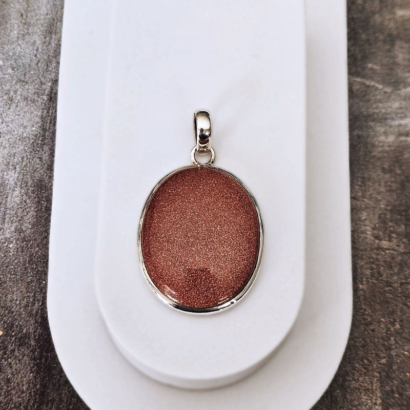 An oval pendant with a shimmering coppery rose gold glitter center, set in a polished silver bezel and bail, displayed on a white card over a dark textured surface.