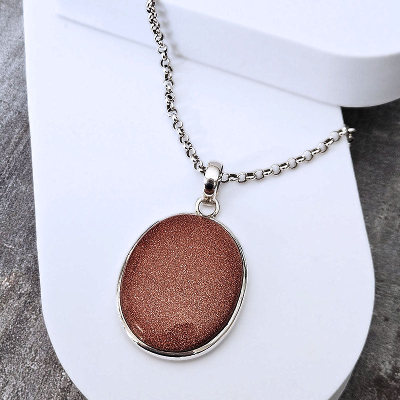 An oval pendant necklace with a shimmering copper-brown center and a silver-tone chain, displayed on a white curved stand.