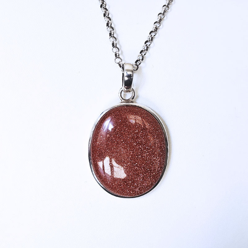 An oval pendant necklace featuring a sparkling reddish-brown stone with fine gold-like shimmer, set in a silver-tone bezel on a silver chain, shown against a plain white background.