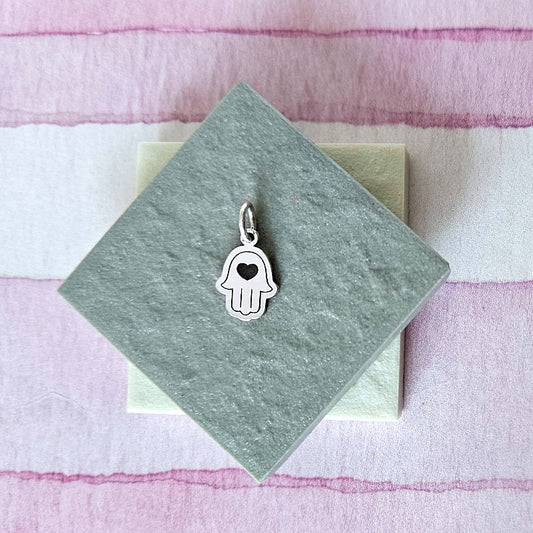 Silver hamsa hand charm on a green card with a striped pink and white background