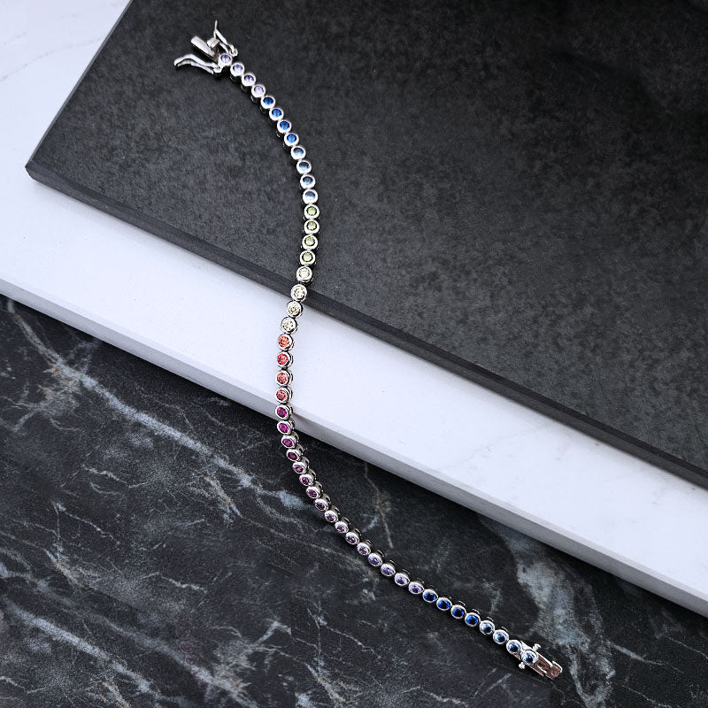 A silver-toned tennis bracelet with a gradient of colored gemstones, laid out diagonally on a black textured slate card over a white marble slab.
