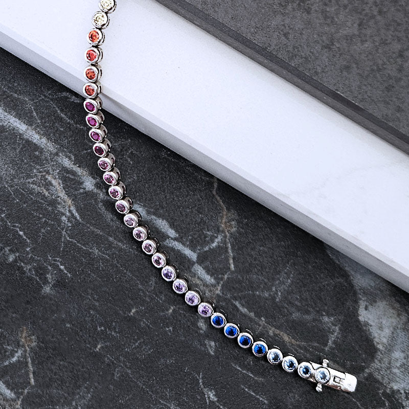 A silver tennis bracelet with alternating colored gemstone beads in red, orange, pink, purple, and blue, displayed on a dark marble surface next to a white book.