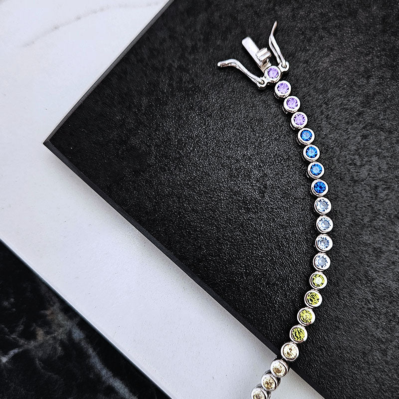 A silver tennis bracelet with a gradient of colored gemstones (purple, blue, white, yellow, green) displayed on a black textured surface.